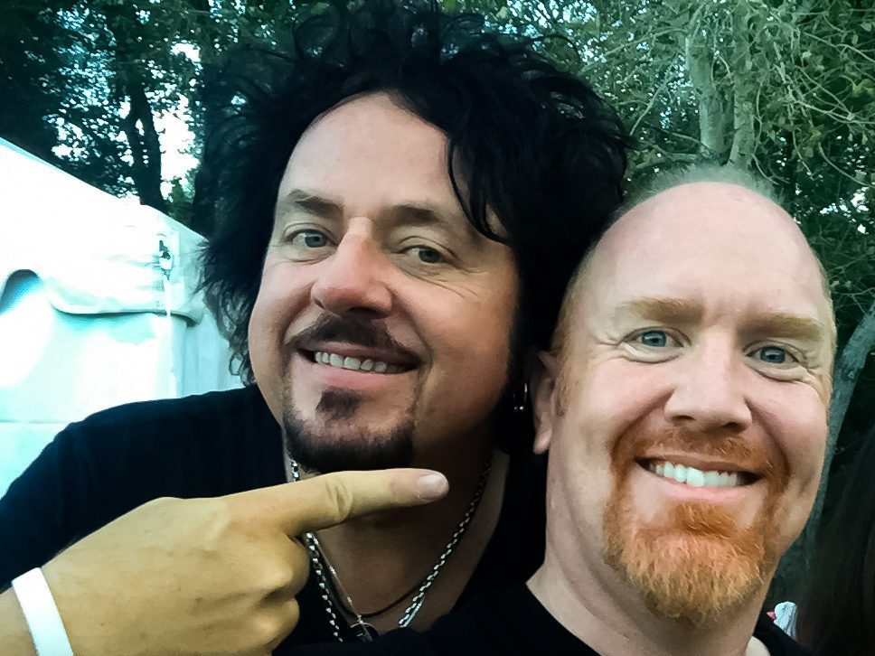 Two men smiling outdoors, one pointing.