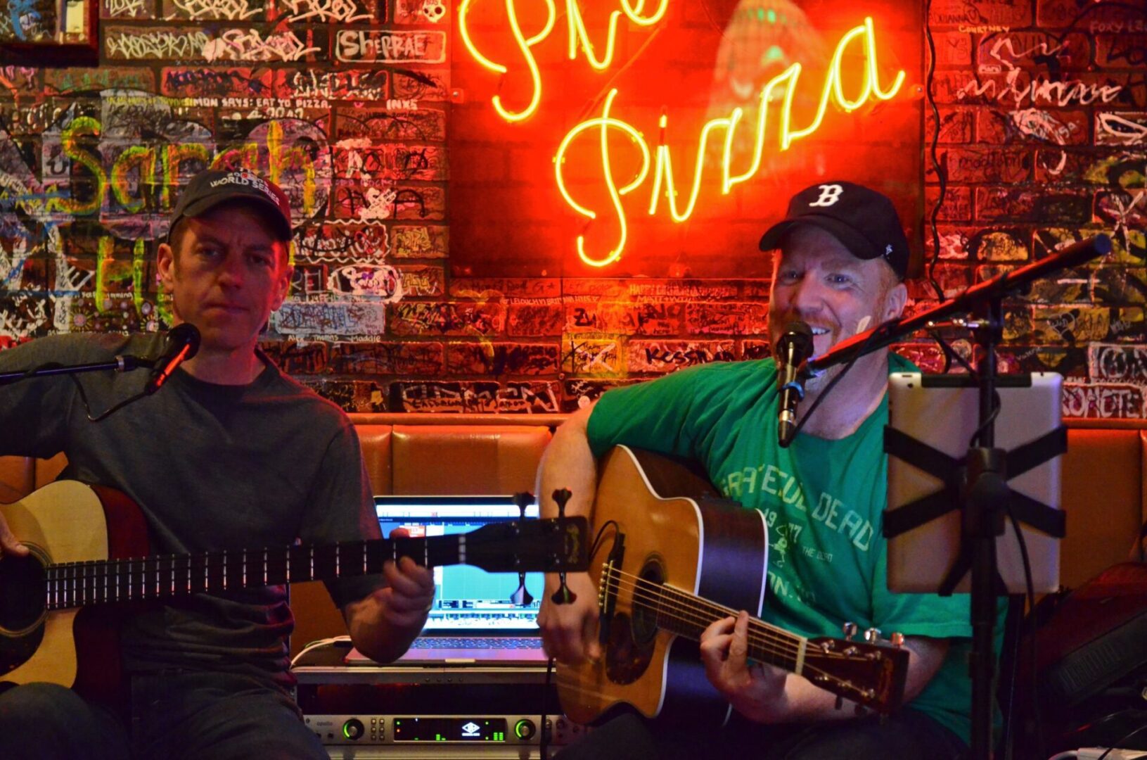 Two men are playing guitar in front of a neon sign.